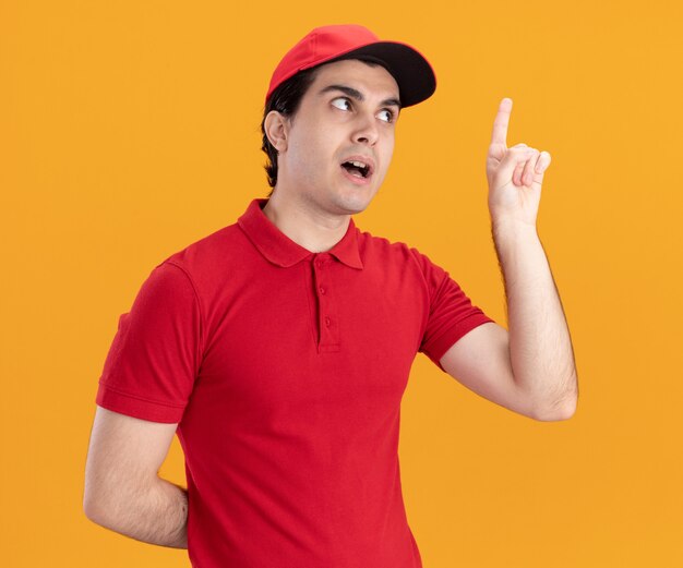 Impresionado joven repartidor caucásico en uniforme azul y gorra manteniendo la mano detrás de la espalda apuntando hacia arriba mirando al lado