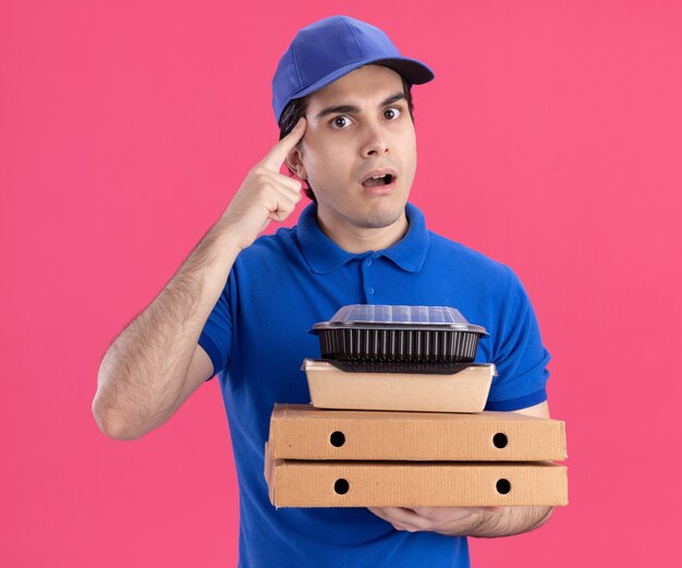 Impresionado joven repartidor caucásico en uniforme azul y gorra sosteniendo paquetes de pizza con contenedor de comida y paquete de comida de papel en ellos haciendo gesto de pensar aislado en la pared rosa