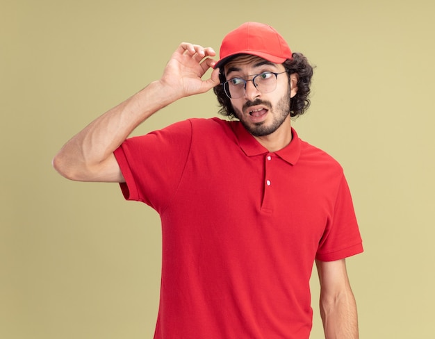 Foto gratuita impresionado joven repartidor caucásico en uniforme rojo y gorra con gafas agarrando gorra mirando al lado