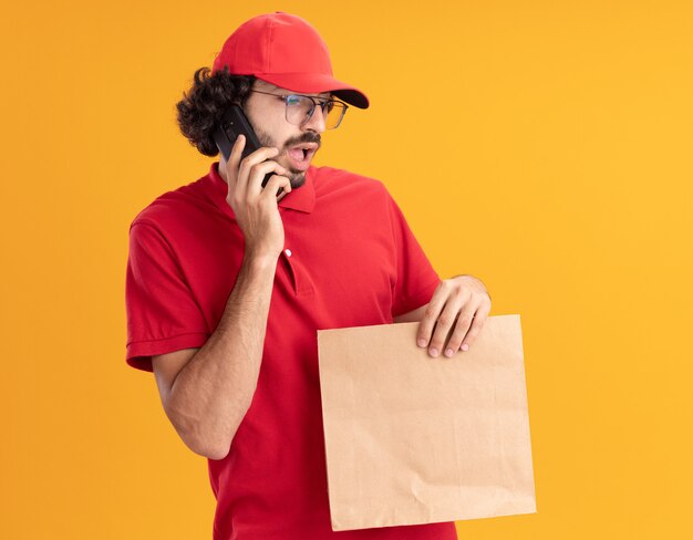 Impresionado joven repartidor caucásico en uniforme rojo y gorra con gafas sosteniendo el paquete de papel hablando por teléfono mirando hacia abajo