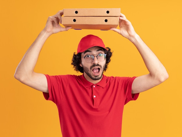 Foto gratuita impresionado joven repartidor caucásico en uniforme rojo y gorra con gafas sosteniendo paquetes de pizza por encima de la cabeza