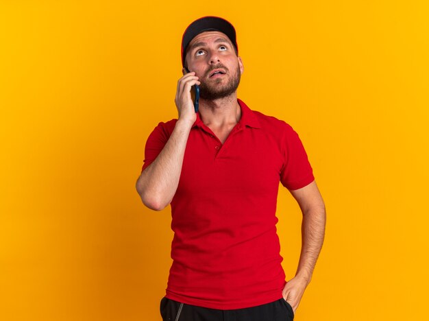 Impresionado joven repartidor caucásico en uniforme rojo y gorra manteniendo la mano en el bolsillo hablando por teléfono mirando hacia arriba aislado en la pared naranja con espacio de copia