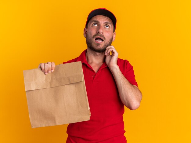 Impresionado joven repartidor caucásico en uniforme rojo y gorra sosteniendo el paquete de papel tocando la cara mirando hacia arriba aislado en la pared naranja con espacio de copia