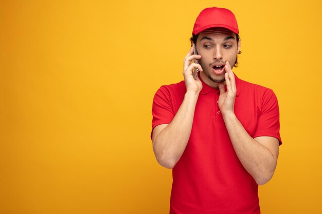 Impresionado joven repartidor con uniforme y gorra manteniendo la mano cerca de la boca mirando al lado susurrando por teléfono aislado en fondo amarillo con espacio para copiar