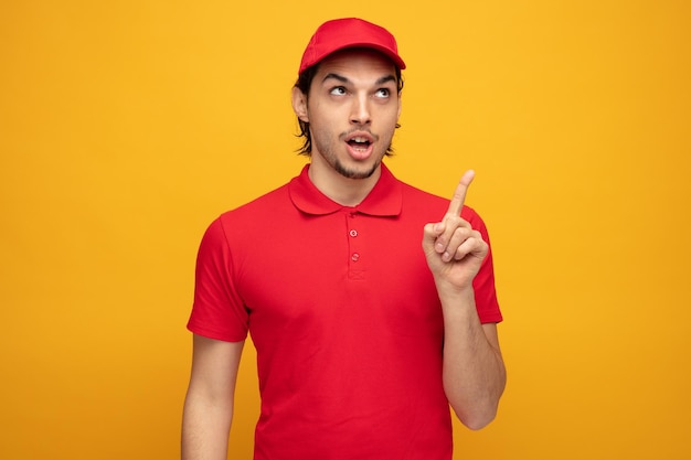 Impresionado joven repartidor con uniforme y gorra mirando y señalando aislado sobre fondo amarillo.