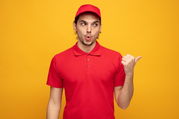 Impresionado joven repartidor con uniforme y gorra mirando y señalando a un lado aislado de fondo amarillo