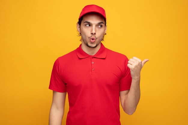 Impresionado joven repartidor con uniforme y gorra mirando y señalando a un lado aislado de fondo amarillo