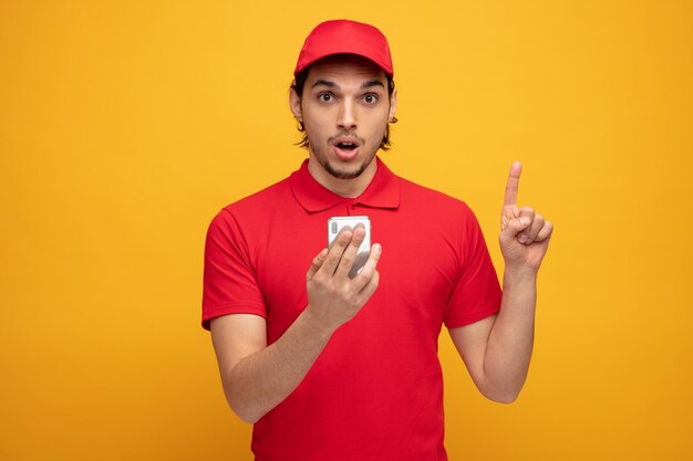 Impresionado joven repartidor vestido con uniforme y gorra sosteniendo un teléfono móvil mirando a la cámara apuntando hacia arriba aislado en un fondo amarillo