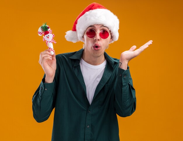 Impresionado joven rubio con gorro de Papá Noel y gafas con adorno de caña dulce mirando a cámara mostrando la mano vacía aislada sobre fondo naranja