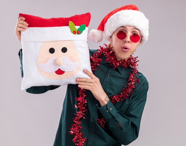 Foto gratuita impresionado joven rubio con gorro de papá noel y gafas con guirnalda de oropel alrededor del cuello sosteniendo la almohada de santa claus mirando a cámara aislada sobre fondo blanco