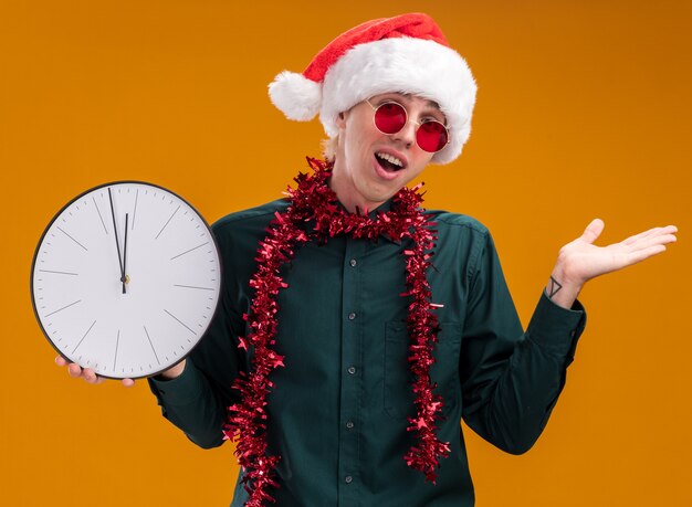 Impresionado joven rubio con gorro de Papá Noel y gafas con guirnalda de oropel alrededor del cuello sosteniendo el reloj mirando a la cámara mostrando la mano vacía aislada sobre fondo naranja