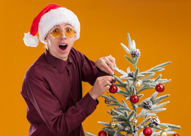 Impresionado joven rubio con gorro de Papá Noel y gafas de pie en la vista de perfil cerca del árbol de Navidad que lo decora con adornos navideños mirando a cámara aislada sobre fondo naranja