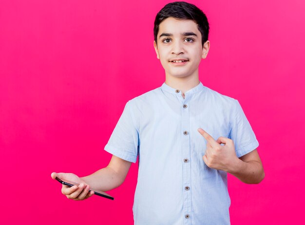 Impresionado joven sosteniendo el teléfono móvil mirando al frente apuntando al lado aislado en la pared rosa