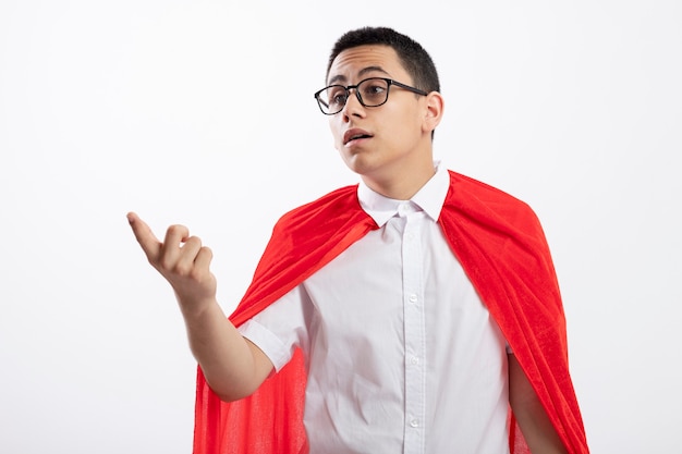 Impresionado joven superhéroe en capa roja con gafas mirando y apuntando al lado aislado sobre fondo blanco.