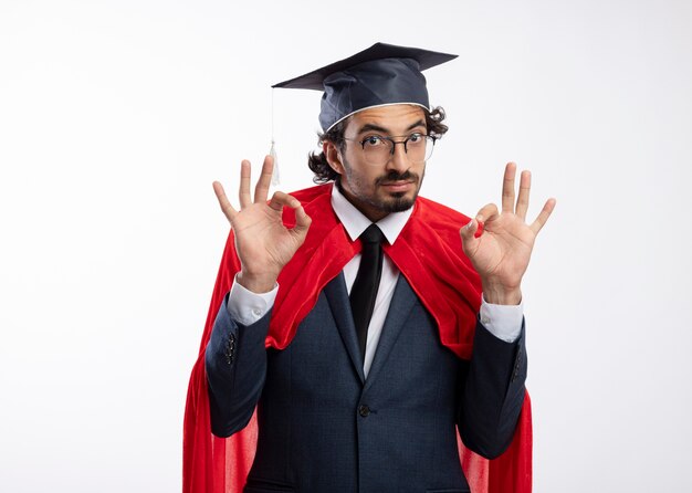 Impresionado joven superhéroe caucásico en gafas ópticas con traje con capa roja y gorro de graduación gestos ok signo de mano con las dos manos