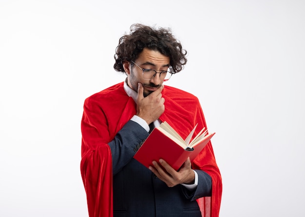 Impresionado joven superhéroe caucásico en gafas ópticas con traje con manto rojo pone la mano en la barbilla y lee el libro