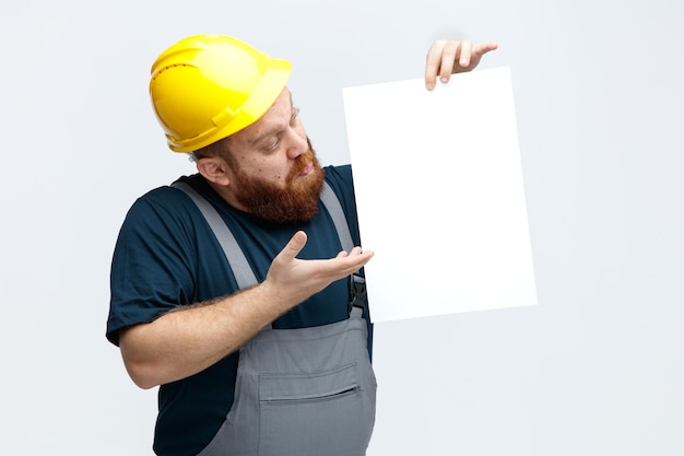 Impresionado joven trabajador de la construcción con casco de seguridad y uniforme mostrando papel a la cámara apuntando al papel con la mano mirándolo aislado en fondo blanco
