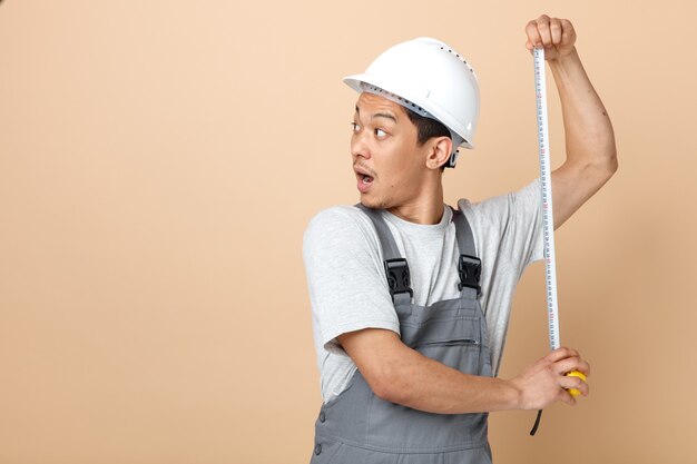 Impresionado joven trabajador de la construcción con casco de seguridad y uniforme sosteniendo medidor de cinta mirando al lado