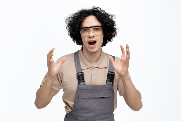 Impresionado joven trabajador de la construcción vistiendo uniforme y gafas de seguridad mirando a la cámara manteniendo las manos en el aire aislado sobre fondo blanco.