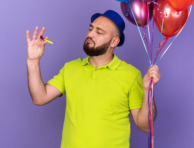 Impresionado joven vestido con gorro de fiesta sosteniendo globos y mirando el silbato en su mano