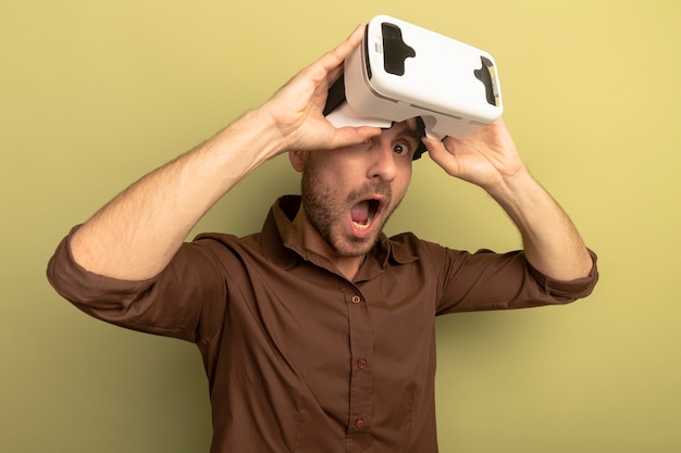 Impresionado joven vistiendo auriculares vr agarrándolo mirando al frente aislado en la pared verde oliva