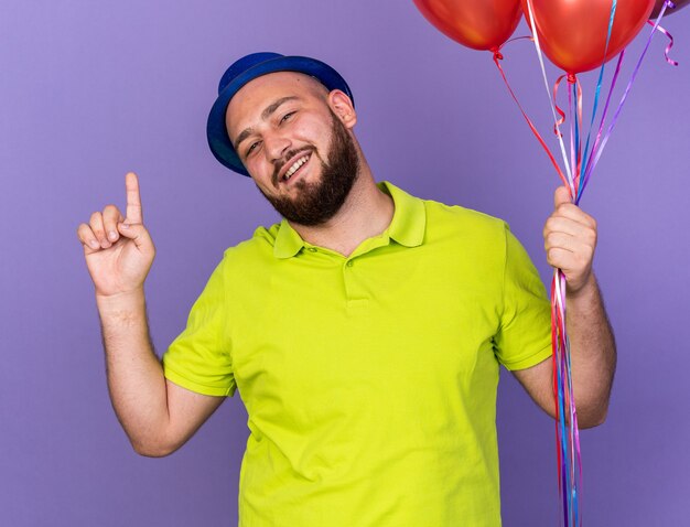 Impresionado joven vistiendo gorro de fiesta sosteniendo globos apunta hacia arriba aislado en la pared azul