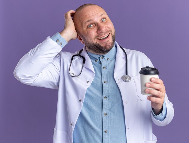 Impresionado médico de mediana edad vistiendo bata médica y estetoscopio tocando la cabeza sosteniendo una taza de café de plástico aislada en la pared púrpura