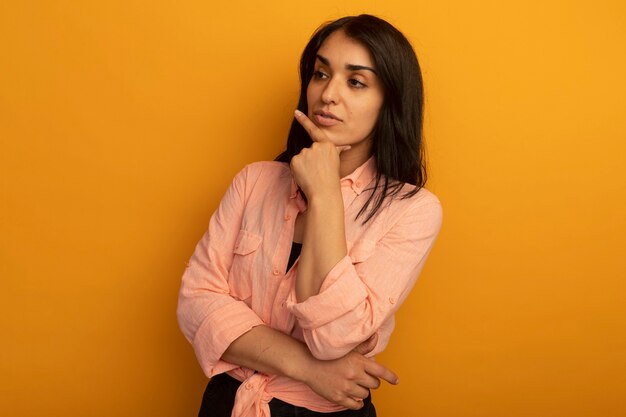 Impresionado mirando al lado hermosa joven vestida con camiseta rosa poniendo la mano en el mentón aislado en amarillo