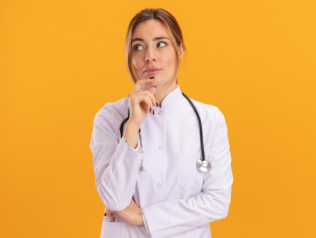 Impresionado mirando al lado joven doctora vistiendo bata médica con estetoscopio agarró la barbilla aislada en la pared amarilla
