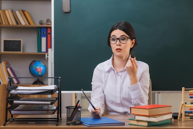 impresionado mirando al lado joven maestra con gafas sosteniendo un puntero sentado en el escritorio con herramientas escolares en el aula