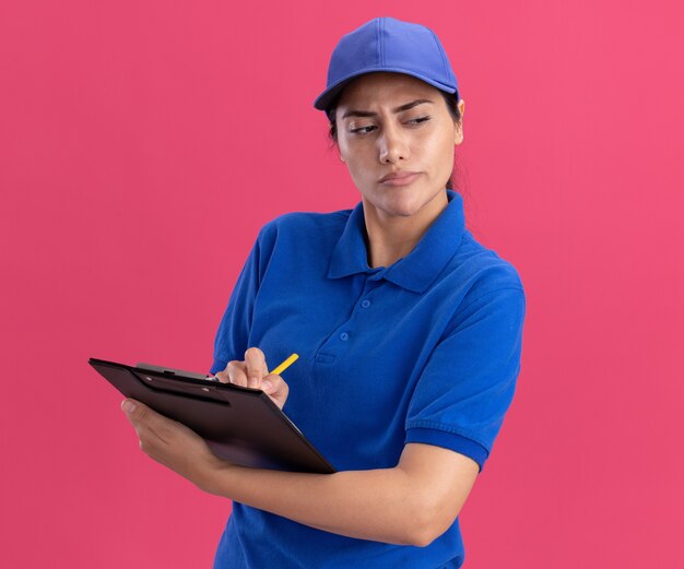 Impresionado mirando al lado joven repartidora vistiendo uniforme con gorra escribiendo algo en el portapapeles aislado en la pared rosa