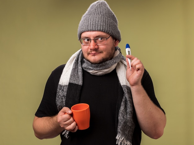 Impresionado mirando al lado masculino enfermo de mediana edad con sombrero de invierno y bufanda sosteniendo una taza de té con termómetro aislado en la pared verde oliva