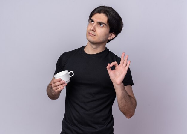 Impresionado mirando hacia arriba chico guapo joven con camiseta negra sosteniendo una taza de café mostrando gesto bien aislado en la pared blanca