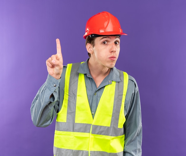 Impresionado mirando a la cámara joven constructor en uniforme apunta hacia arriba