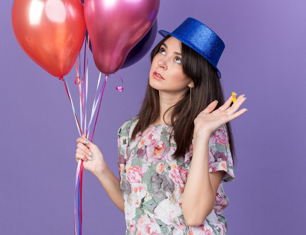 Impresionado mirando lateral hermosa joven mujer vistiendo gorro de fiesta sosteniendo globos extendiendo la mano aislada en la pared azul