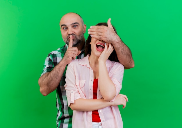 Foto gratuita impresionado pareja adulta hombre cubriendo los ojos de la mujer con la mano gesticulando silencio mujer poniendo la mano en la cara aislada en la pared verde con espacio de copia