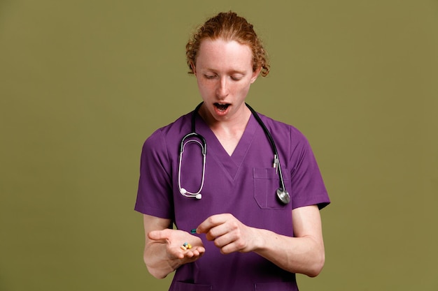 Impresionado sosteniendo pastillas joven médico masculino vistiendo uniforme con estetoscopio aislado sobre fondo verde