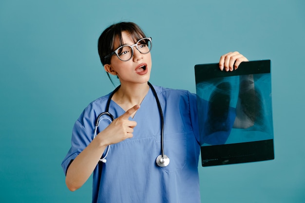 Impresionado sosteniendo rayos X joven doctora vistiendo uniforme fith estetoscopio aislado sobre fondo azul.