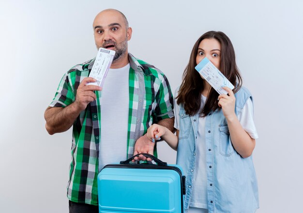 Impresionado viajero adulto pareja hombre sujetando la maleta y tocando la barbilla con billetes de viaje y mirando