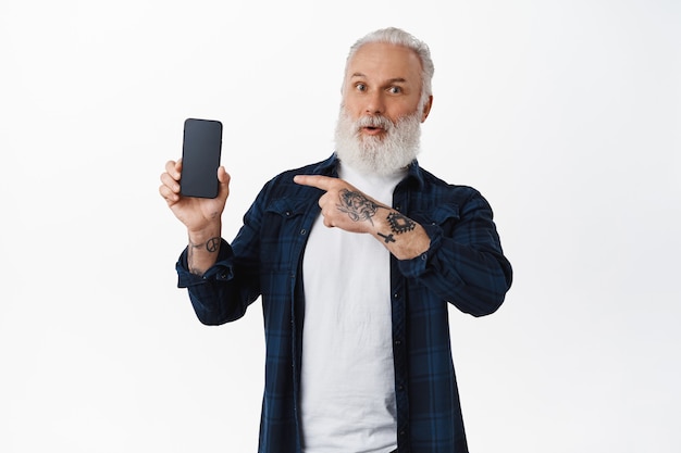 Impresionado viejo tatuado apuntando a la pantalla del teléfono inteligente, mostrando una nueva aplicación móvil increíble, muestra una aplicación, de pie sobre una pared blanca