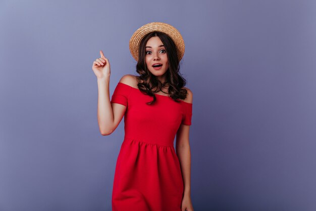 Impresionante chica con cabello ondulado posando en la pared púrpura con expresión de cara de sorpresa. Retrato interior de dama de buen humor con vestido rojo y sombrero de paja.