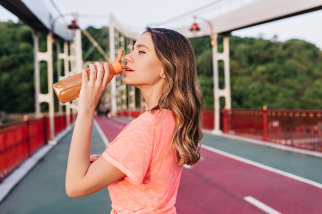 Impresionante chica en camiseta rosa agua potable después del entrenamiento. Encantadora modelo femenina con cabello brillante pasar tiempo en el estadio.