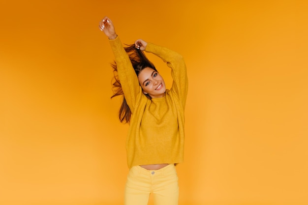 Impresionante chica caucásica en pantalones amarillos bailando divertido en el estudio Adorable modelo femenino agitando su cabello sobre fondo naranja