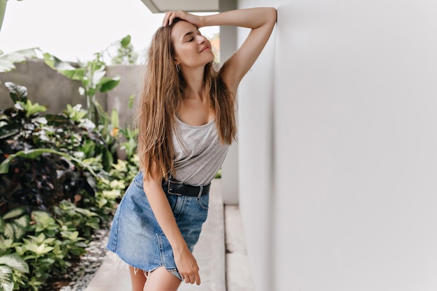 Impresionante chica delgada posando cerca de plantas con expresión facial inspirada. Magnífica dama en falda de mezclilla retro escalofriante.