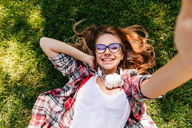 Impresionante chica europea tumbado en la hierba y riendo. Mujer joven agradable que presenta en el parque con sonrisa alegre.