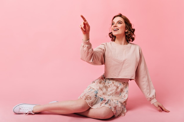 Impresionante chica en falda sentada sobre fondo rosa con sonrisa. Mujer joven rizada inspirada que señala con el dedo.