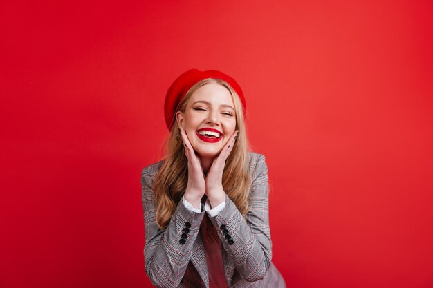 Impresionante chica francesa posando en la pared roja. Alegre joven rubia sonriendo