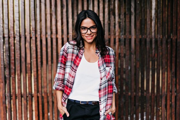 Impresionante chica con gafas de pie en pose de confianza y sonriendo. Disparo al aire libre de dama latina posando con la mano en el bolsillo en la pared de madera.