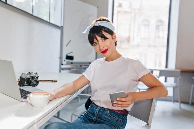 Impresionante chica de pelo negro con teléfono en mano tomando café y descansando en casa