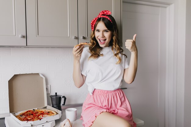 Impresionante chica con pelo rizado brillante pasando la mañana con una taza de té. Riendo a una mujer bonita comiendo pizza.